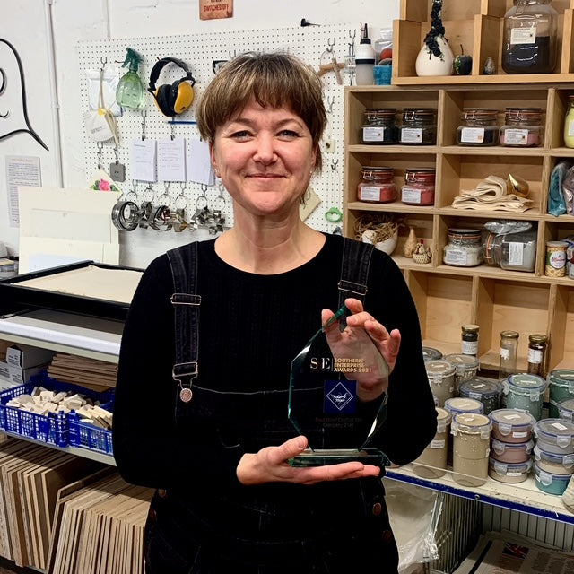 Portrait image of Mel Chambers holding the SME Southern Enterprise Award trophy in her workshop, surrounded by glass jars and other workshop items in the background.